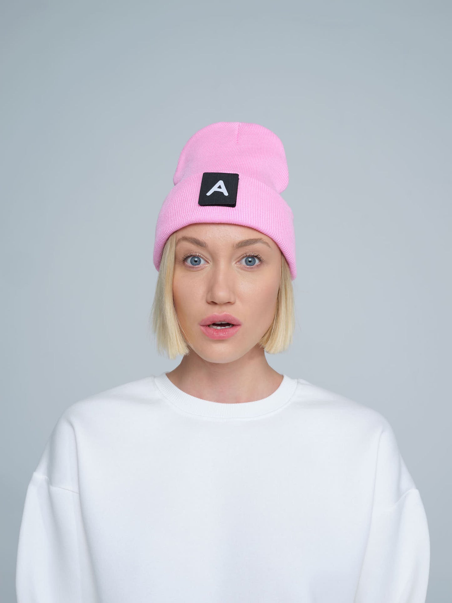 A person wearing a pink beanie with a design on top and the letter 'A' on it, paired with a white crew neck sweater.