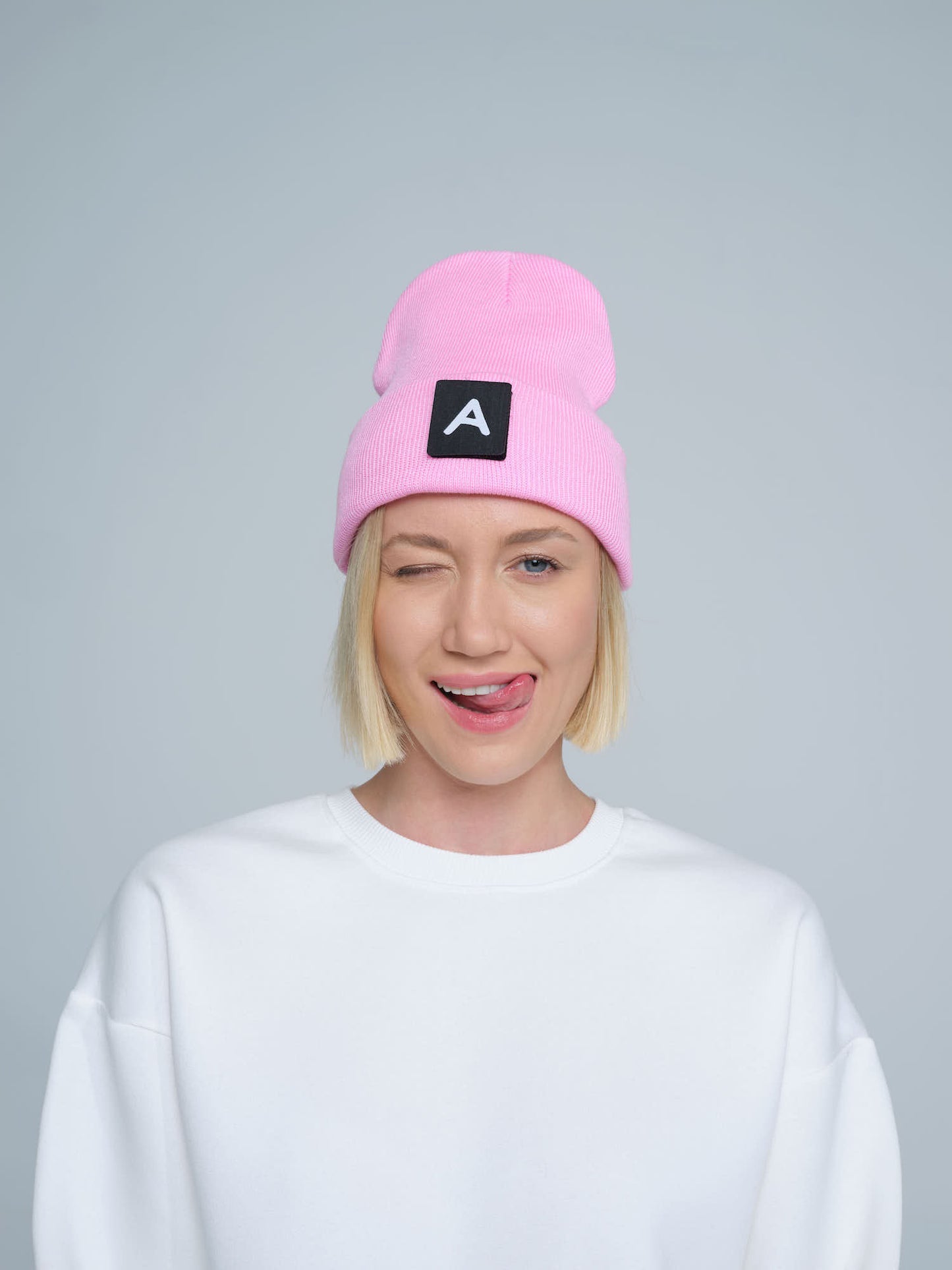 A person wearing a pink beanie with a design on top and the letter 'A' on it, paired with a white crew neck sweater.