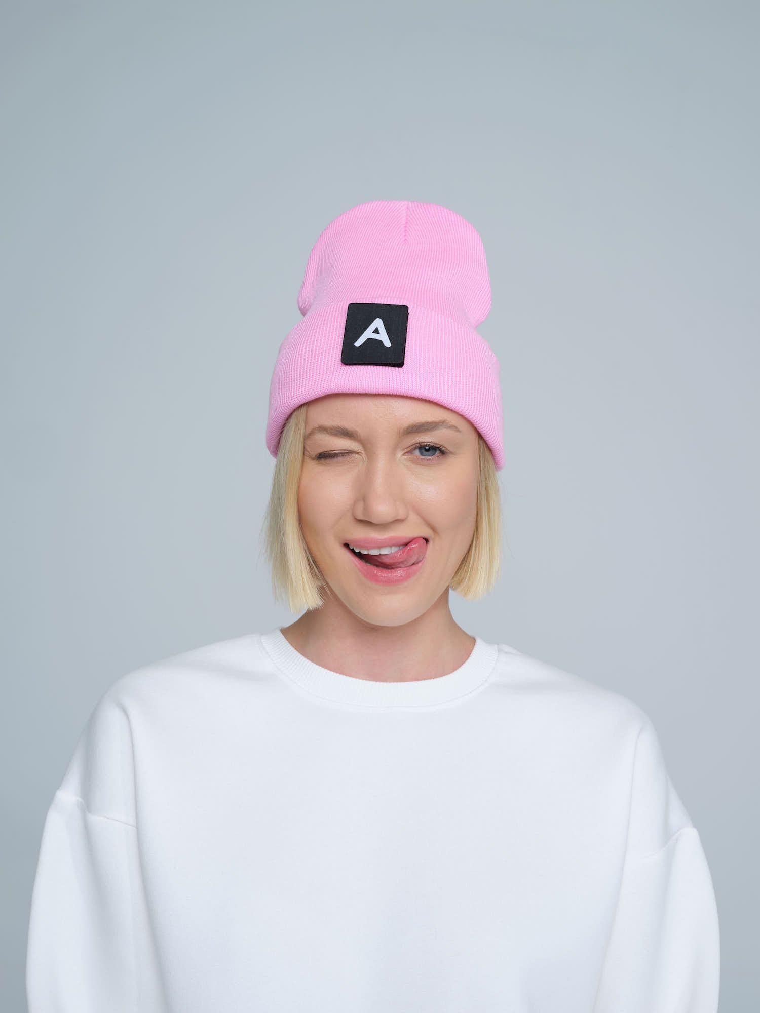 A person wearing a pink beanie with a design on top and the letter 'A' on it, paired with a white crew neck sweater.