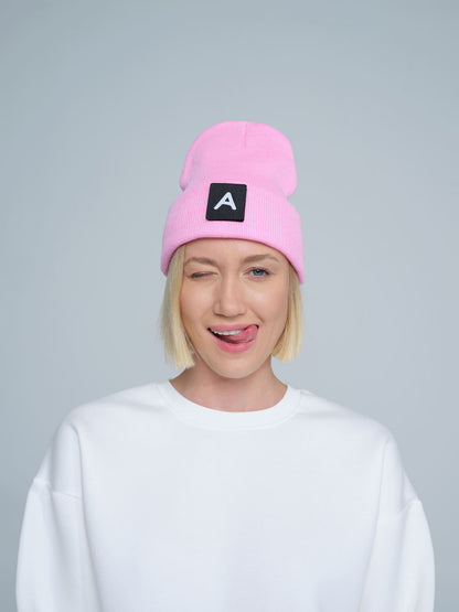A person wearing a pink beanie with a design on top and the letter 'A' on it, paired with a white crew neck sweater.