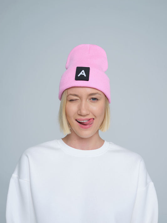 A person wearing a pink beanie with a design on top and the letter 'A' on it, paired with a white crew neck sweater.