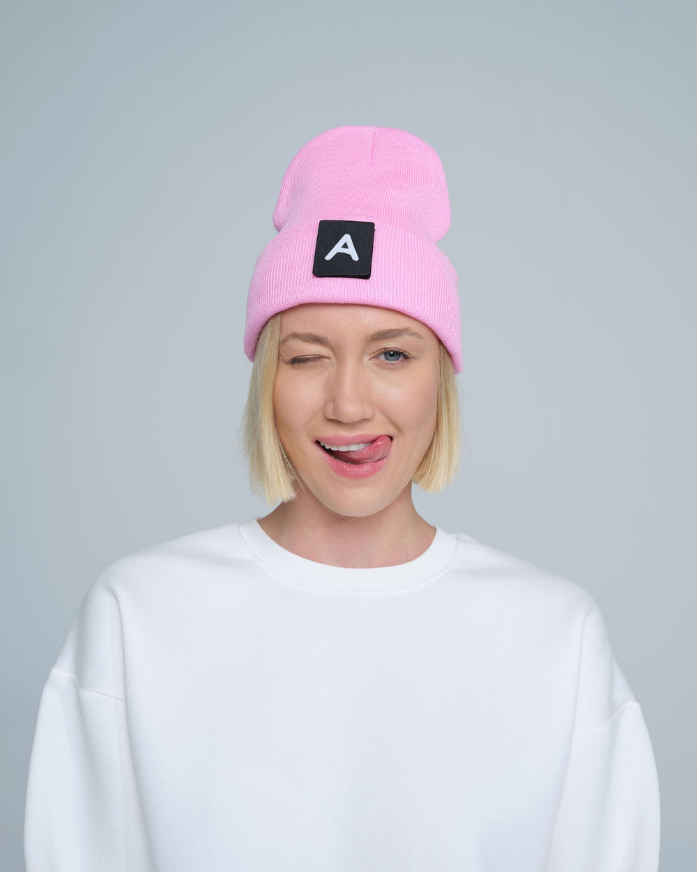 A person wearing a pink beanie with a design on top and the letter 'A' on it, paired with a white crew neck sweater.