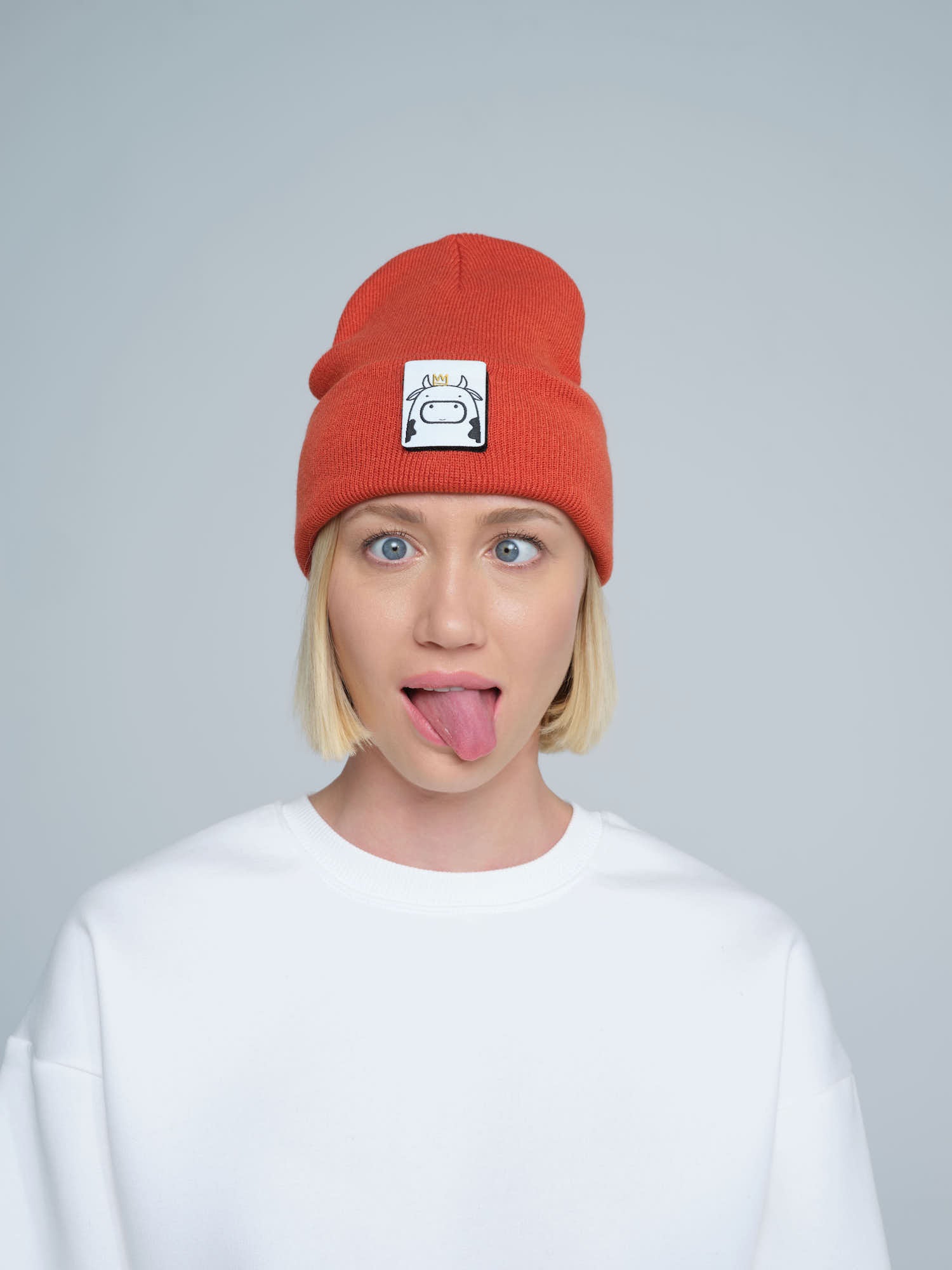 A person wearing a rust orange beanie hat with a cow design patch on the front with a funny face. 