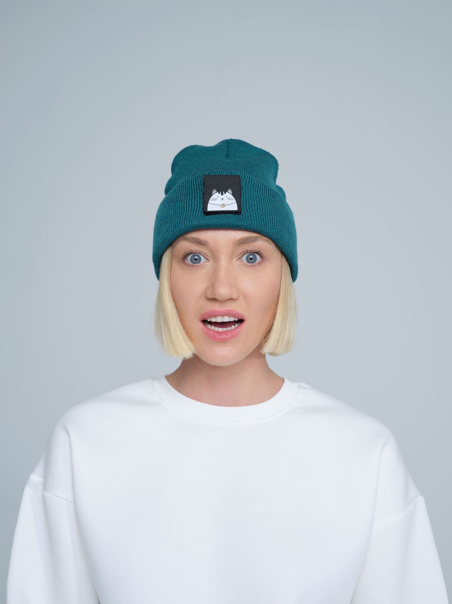 A person wearing a teal green beanie hat with a cat design on the cuff, paired with a white t-shirt.
