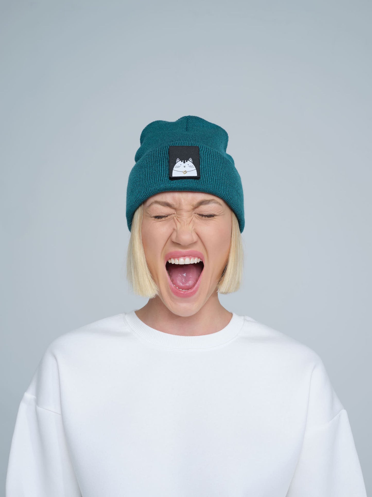 A female wearing a teal green beanie hat with a cat design on the cuff, making a scream face. 