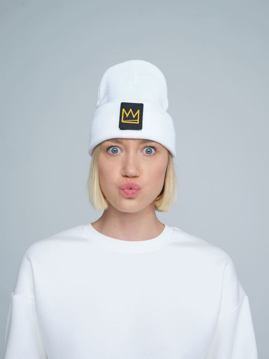 A person wearing a white beanie with a design and a crown symbol on it, standing in a casual pose.