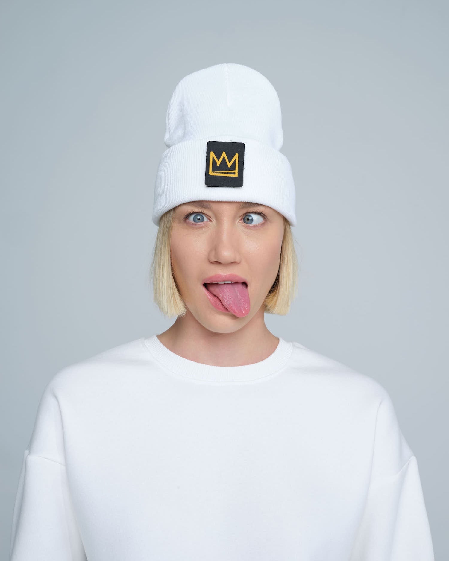 A female wearing a white beanie with a crown symbol on it, standing in a funny pose.