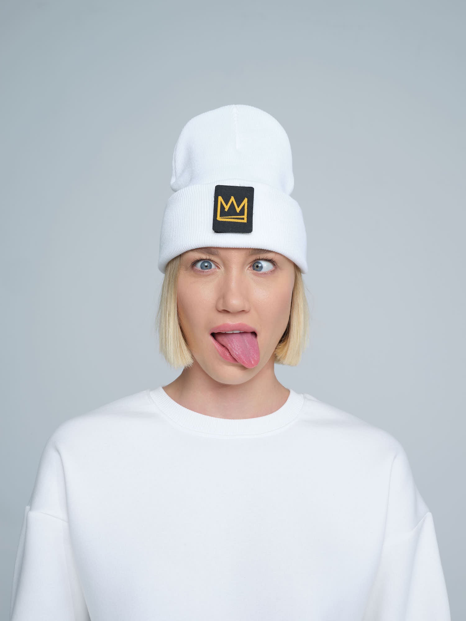 A female wearing a white beanie with a crown symbol on it, standing in a funny pose.